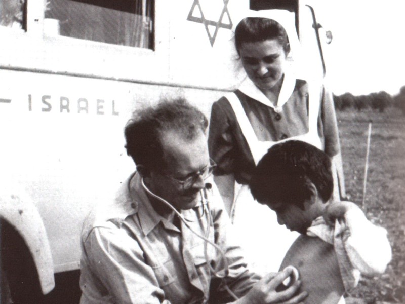 איש צוות רפואי בודק ילד ליד מרפאה ניידת בישראל, ואשת צוות רפואי צופה