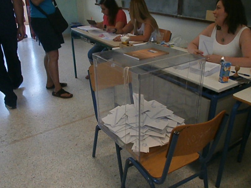 קלפי שקופה מלאה בפתקים ולצדה צוות קלפי ומצביעים במשאל העם היווני על תוכנית החילוץ הכלכלית