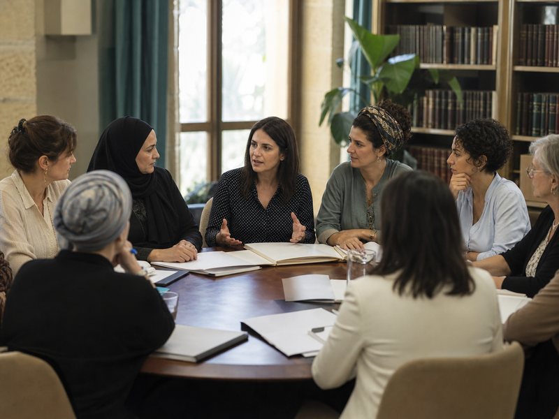 נשים יושבות סביב שולחן דיונים שעליו ספרים פתוחים ותיקיות
