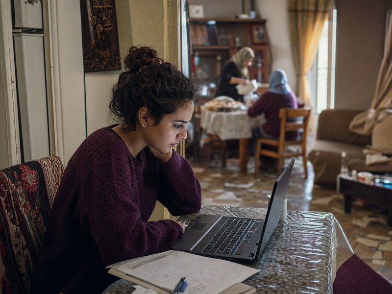 סטודנטית פלסטינית־ערבייה לומדת במחשב נייד בפינת בית משותפת לצד מחברות וחפצי משפחה ללא טקסט קריא