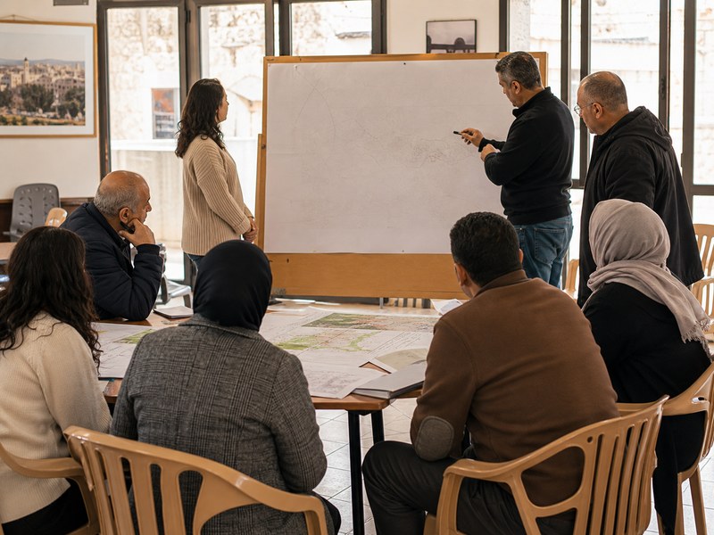 חדר קהילתי ביישוב ערבי שבו תושבים וחוקרים בוחנים מפה ריקה ונתוני בחירות לא קריאים